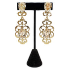 Gold earrings with clear gemstones on a black stand against a white background